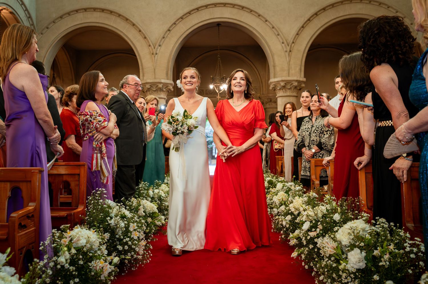 Bride walks down church aisle with woman in red dress, flanked by floral arrangements and wedding guests.