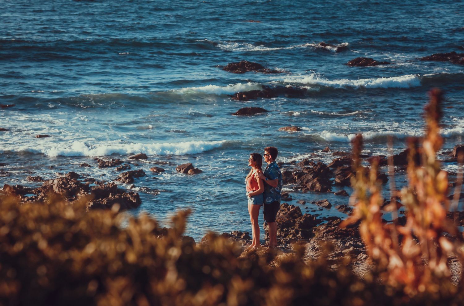 Couple standing close together near rocky shoreline with ocean waves.