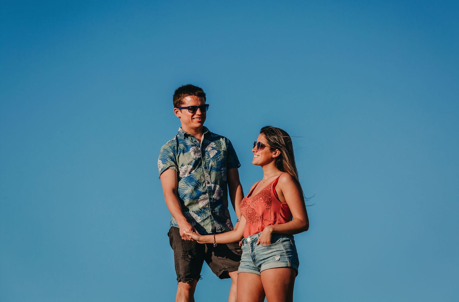 Stylish couple holding hands against bright blue summer sky.