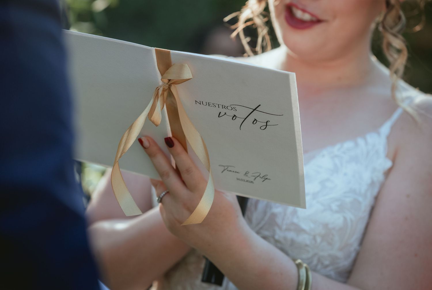 Bride holds elegant white wedding vow booklet tied with gold ribbon during outdoor ceremony.