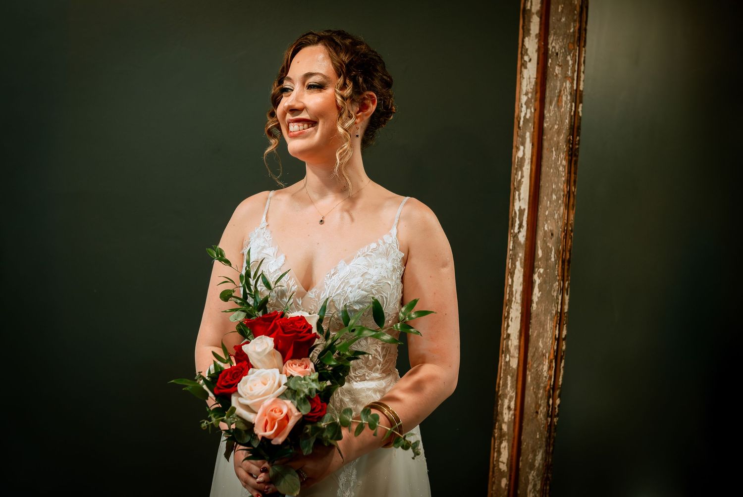 Smiling bride in lace wedding gown holds colorful red and pink rose bouquet beside gold mirror.