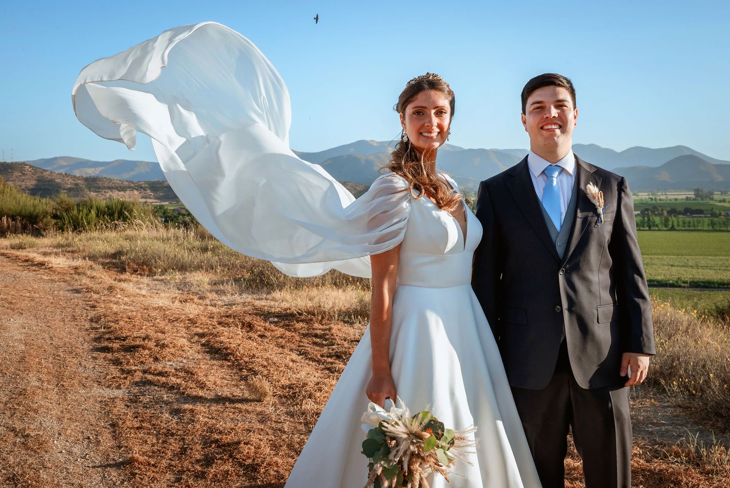 Bride and groom pose outdoors with veil flowing in wind, mountains and green fields in background.