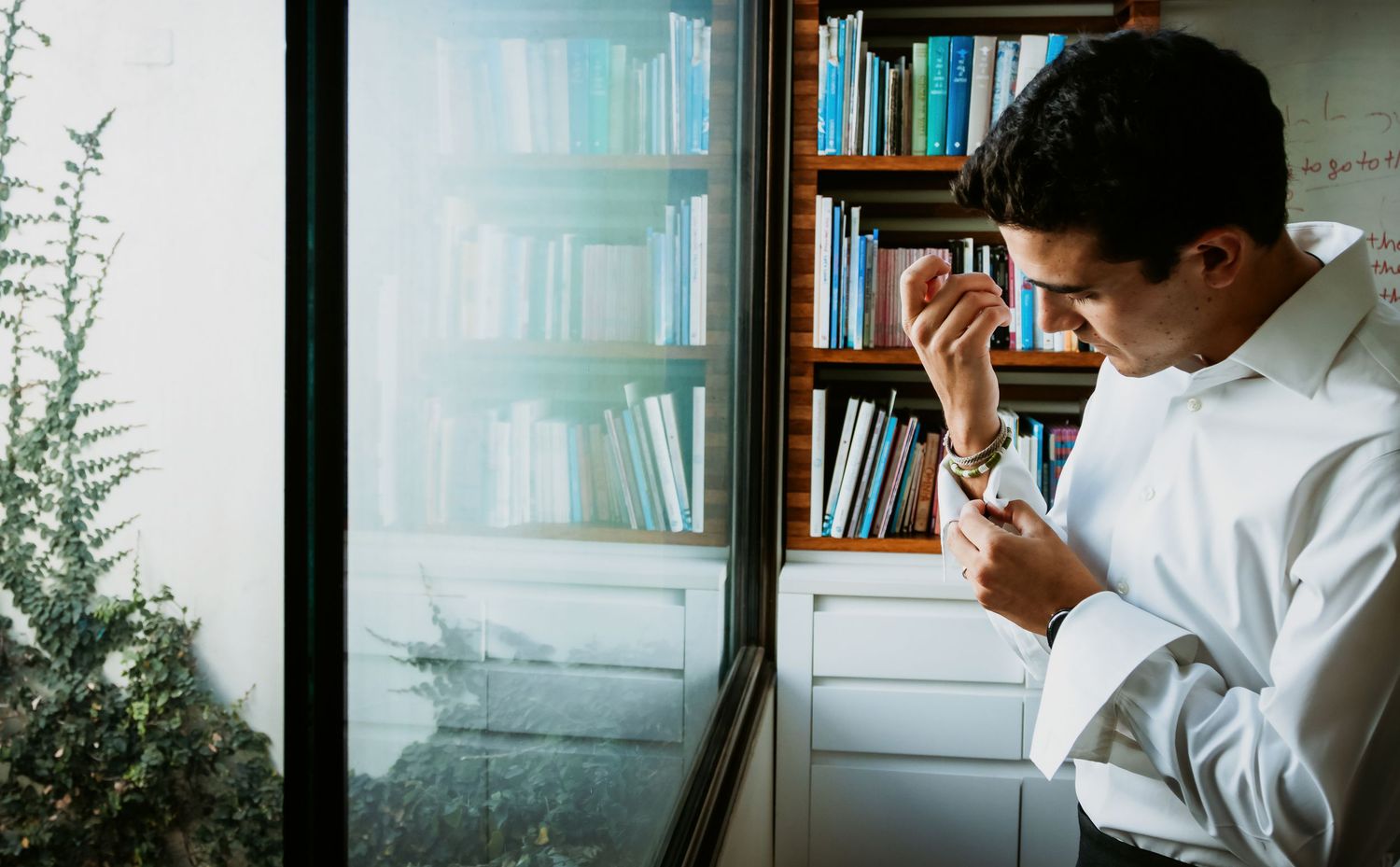 Groom in white shirt adjusting cufflinks by window with bookshelf in background.