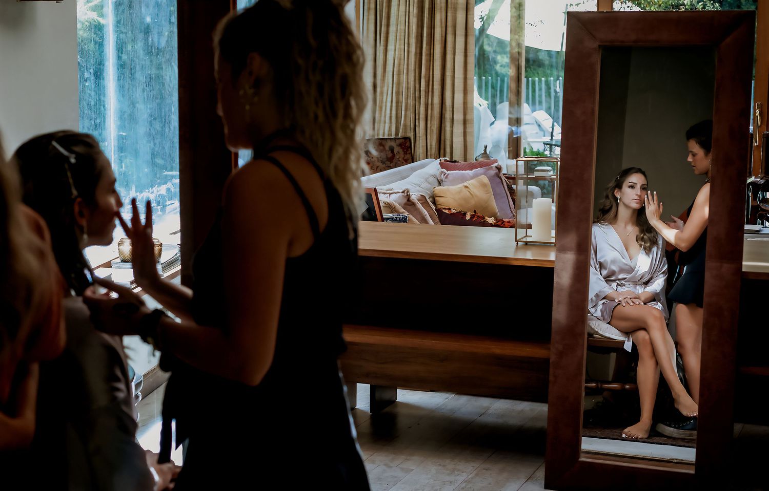 Bride in satin robe sitting before mirror while wedding prep team works on hair and makeup in suite.