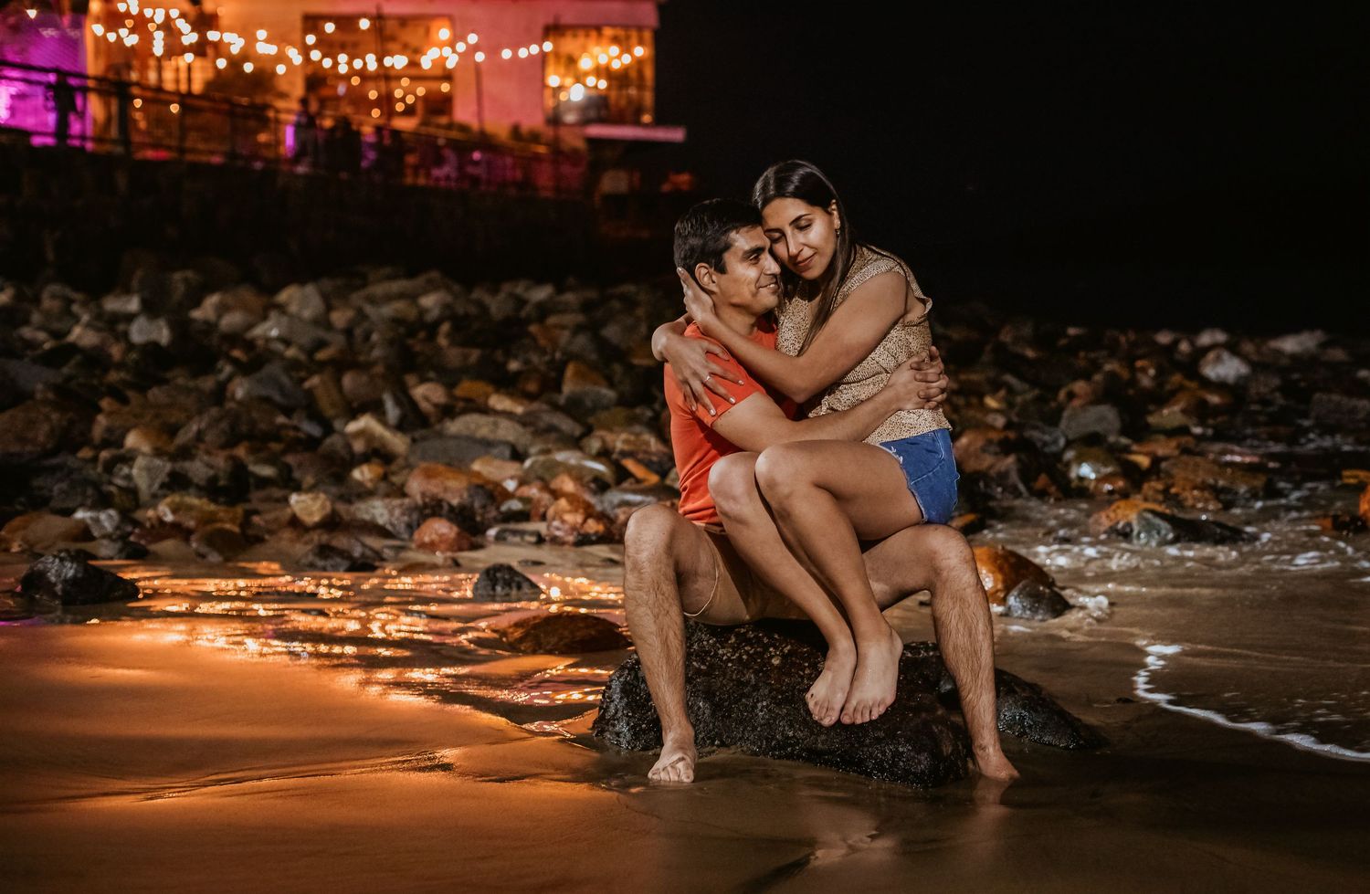 Couple sitting romantically on rocks at a beach at night with warm lights glowing in the background.