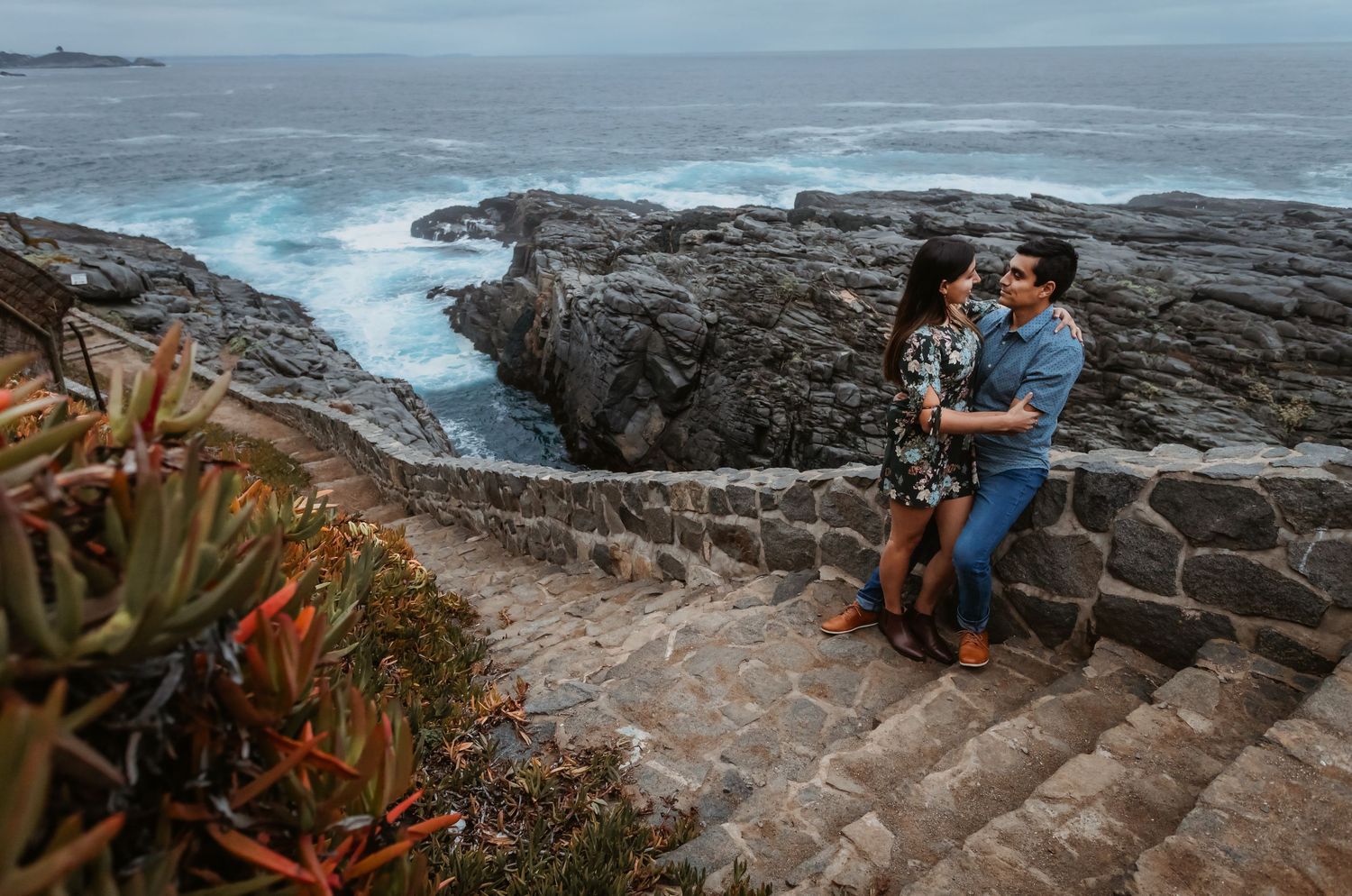 Couple embracing on rocky coastal steps with dramatic ocean waves crashing below.