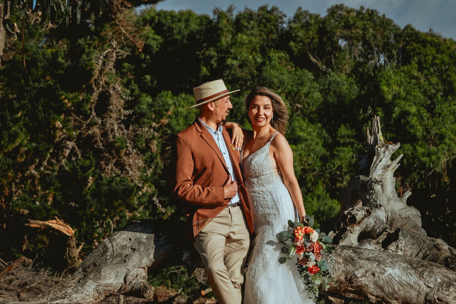 Smiling couple in wedding attire, the groom in a straw hat and rust blazer and the bride in a white lace gown, posing by driftwood with gree