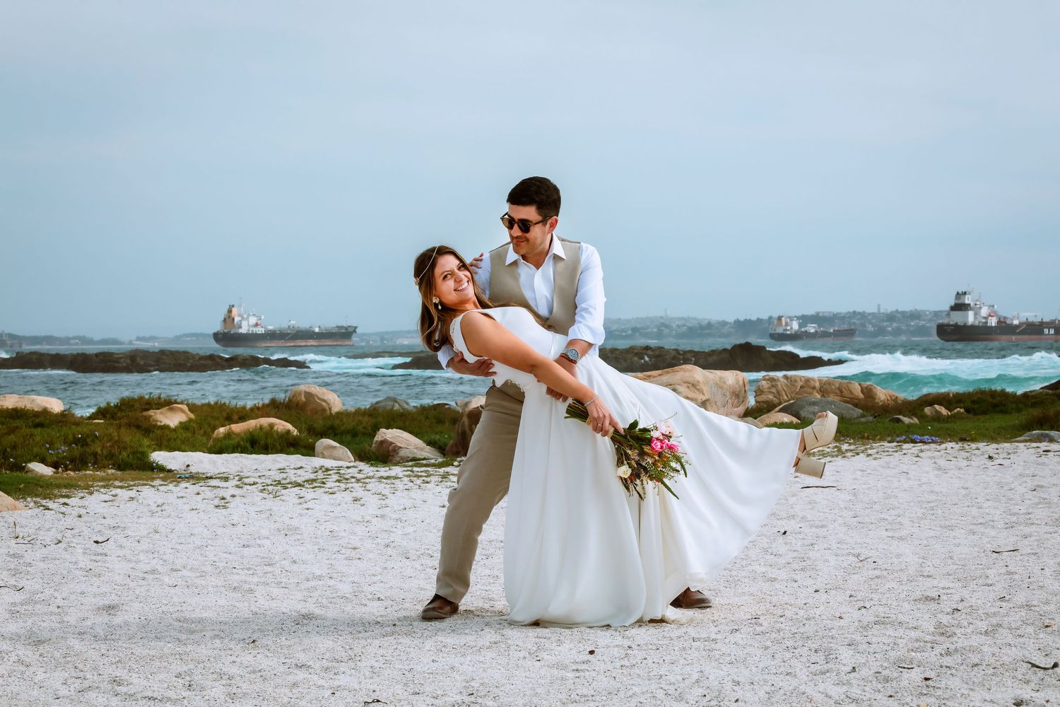 Groom dipping bride in white wedding dress on sandy beach with ocean and ships visible in background.