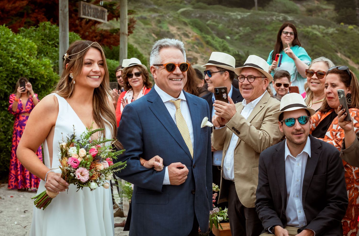 Bride in white dress holding bouquet walks with older man in blue suit at outdoor wedding ceremony.