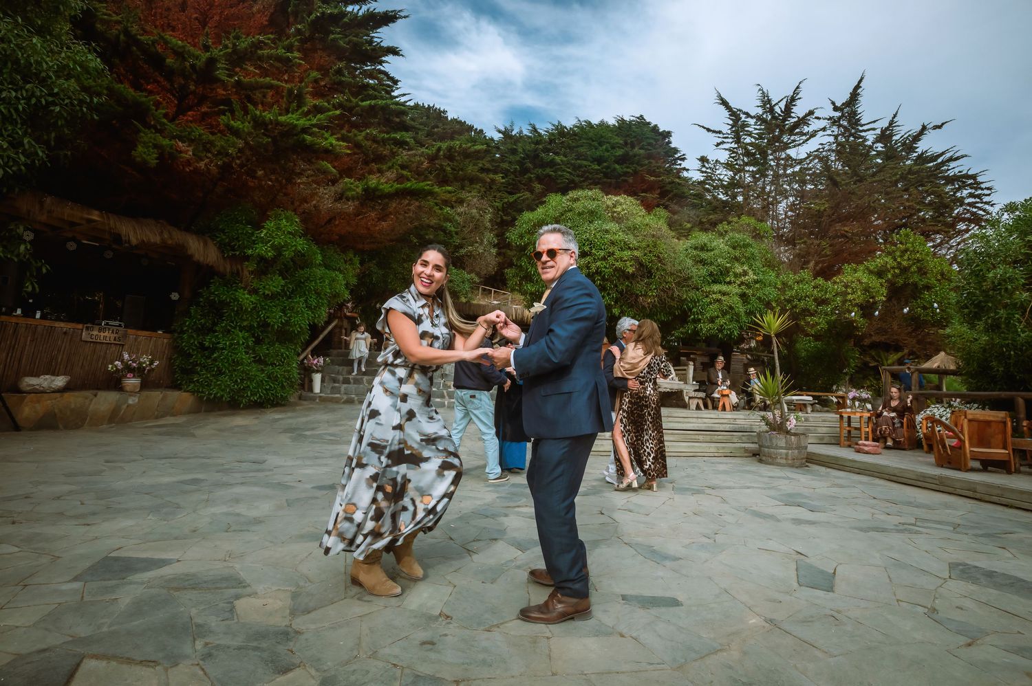 Couple dancing together on a stone patio at an outdoor wedding venue surrounded by lush green trees.