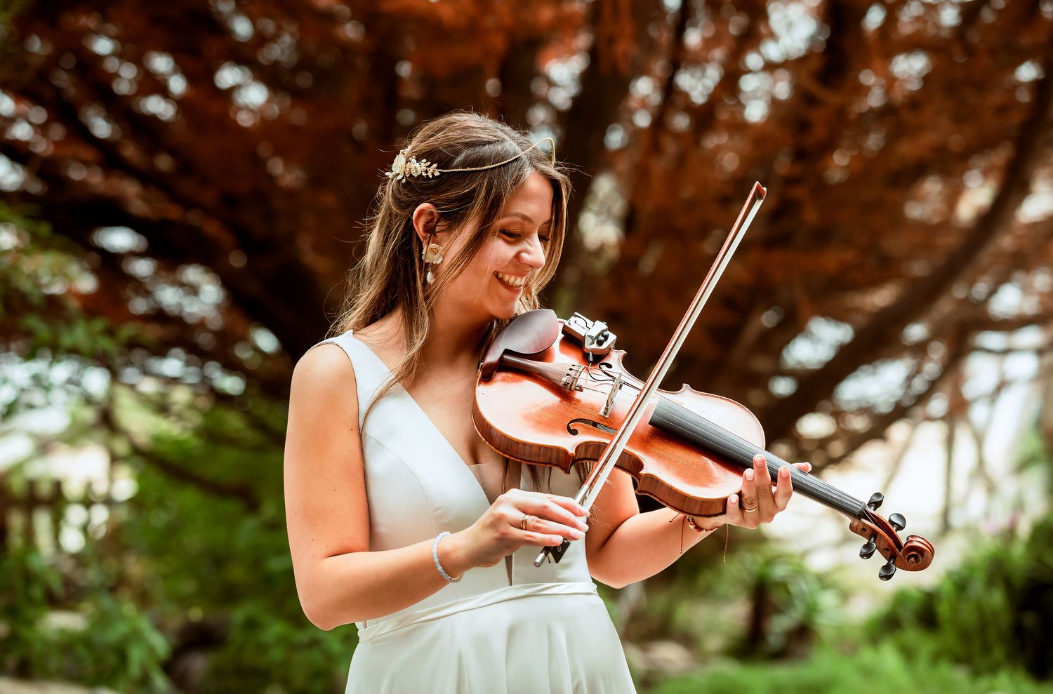 Young woman in white dress plays violin outdoors under a large tree at a wedding celebration.
