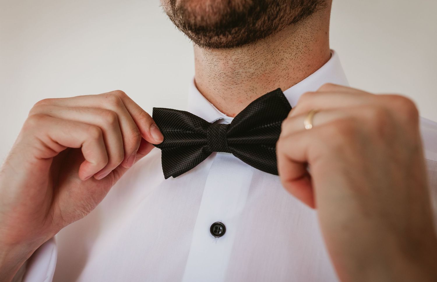 Close-up of groom adjusting black bow tie wearing white tuxedo shirt with wedding ring.