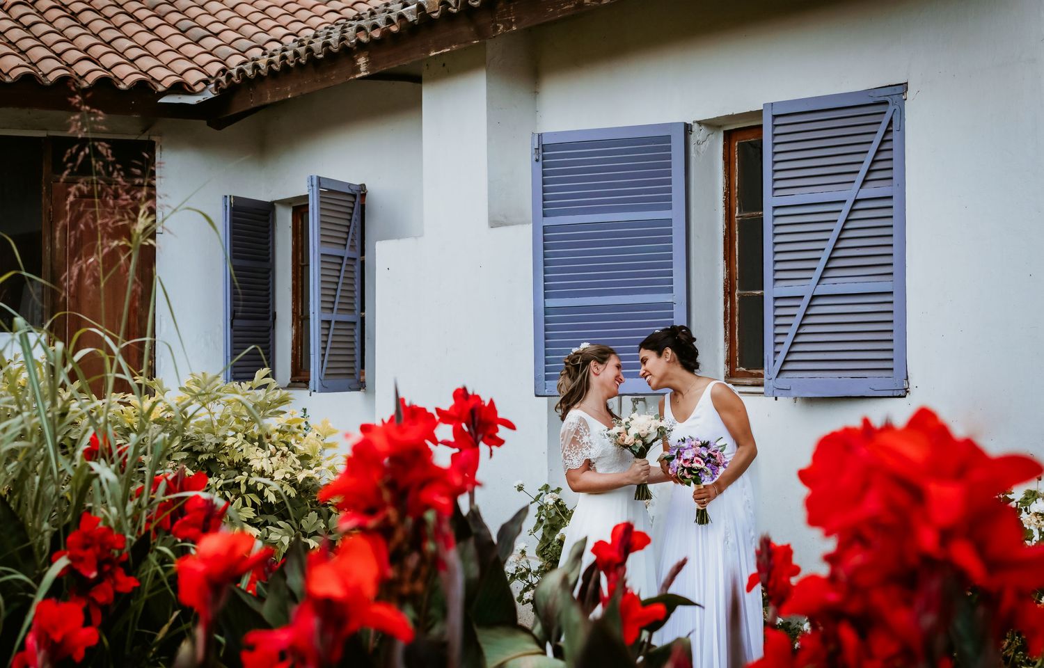 Two brides sharing an intimate moment holding bouquets beside white building with red flowers foreground.