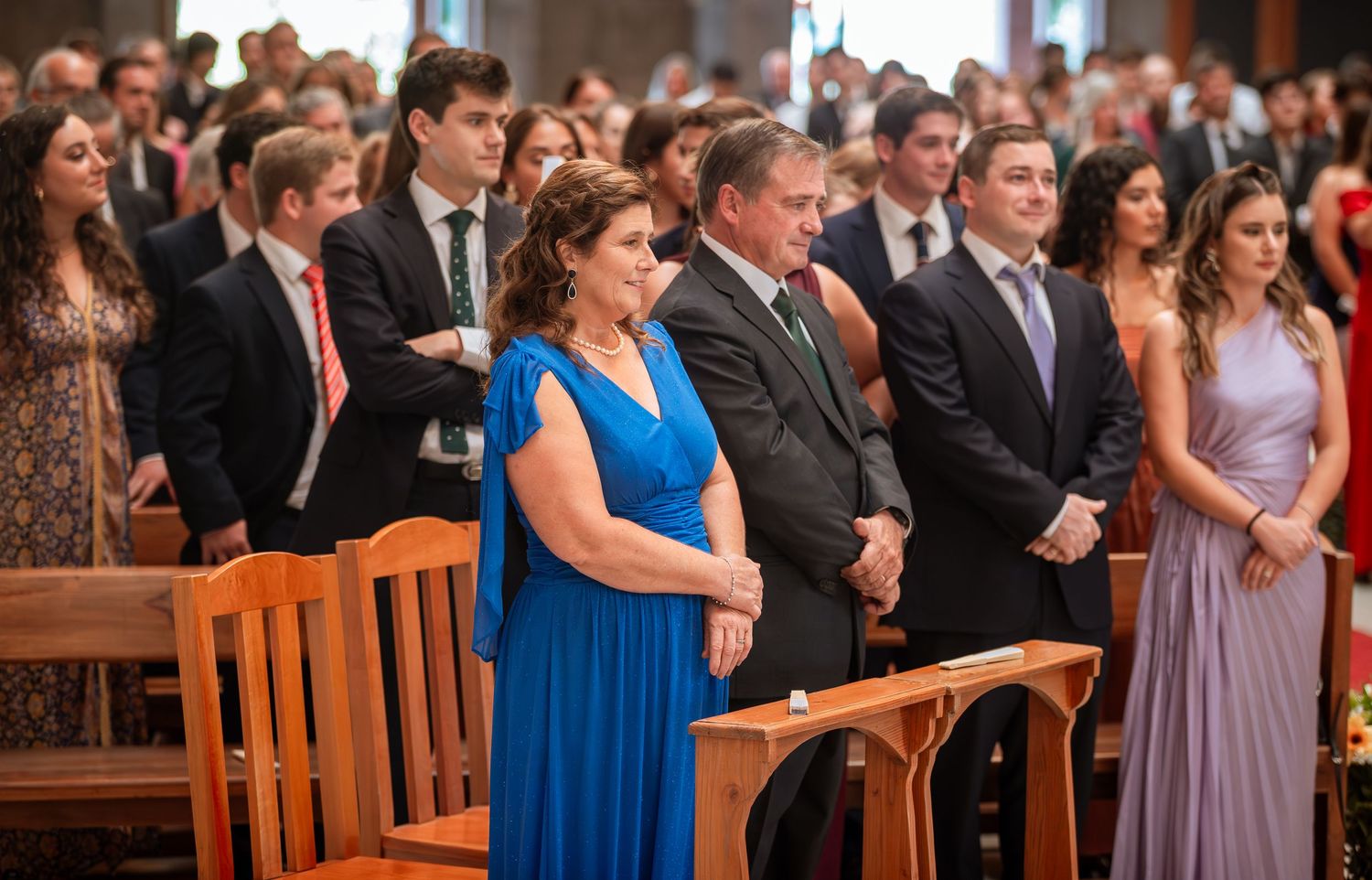 Wedding guests in formal attire stand during ceremony in church with wooden chairs.