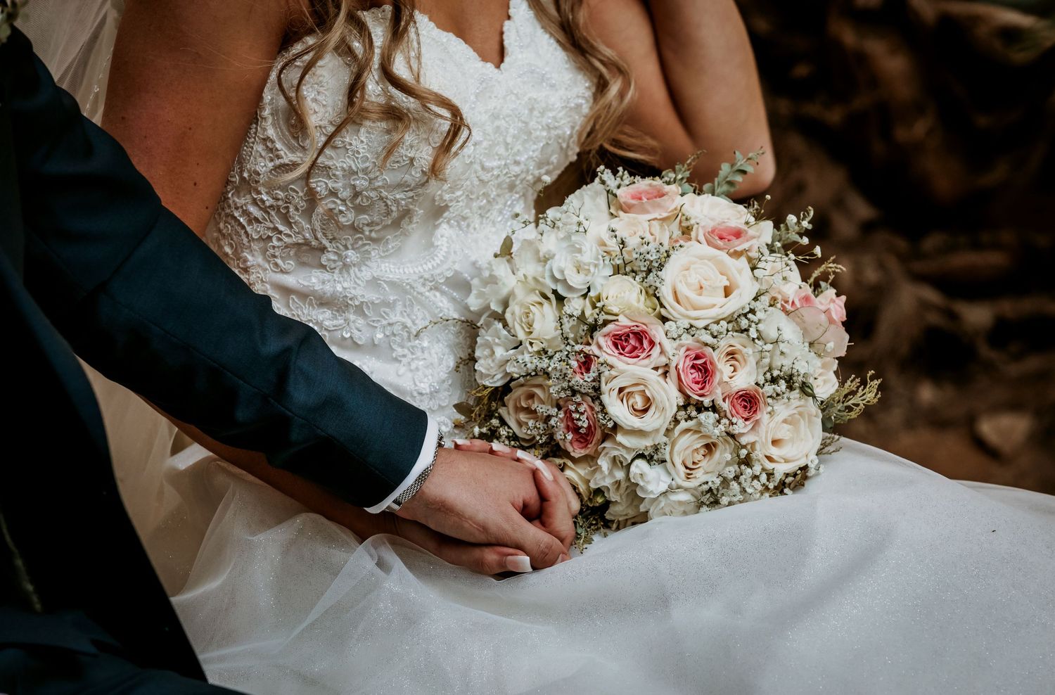 Close-up of bride and groom holding hands with elegant pink and white rose bouquet.