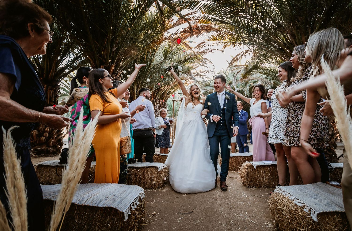 Newlyweds walk joyfully through a celebratory aisle of guests waving dried pampas grass at their wedding.