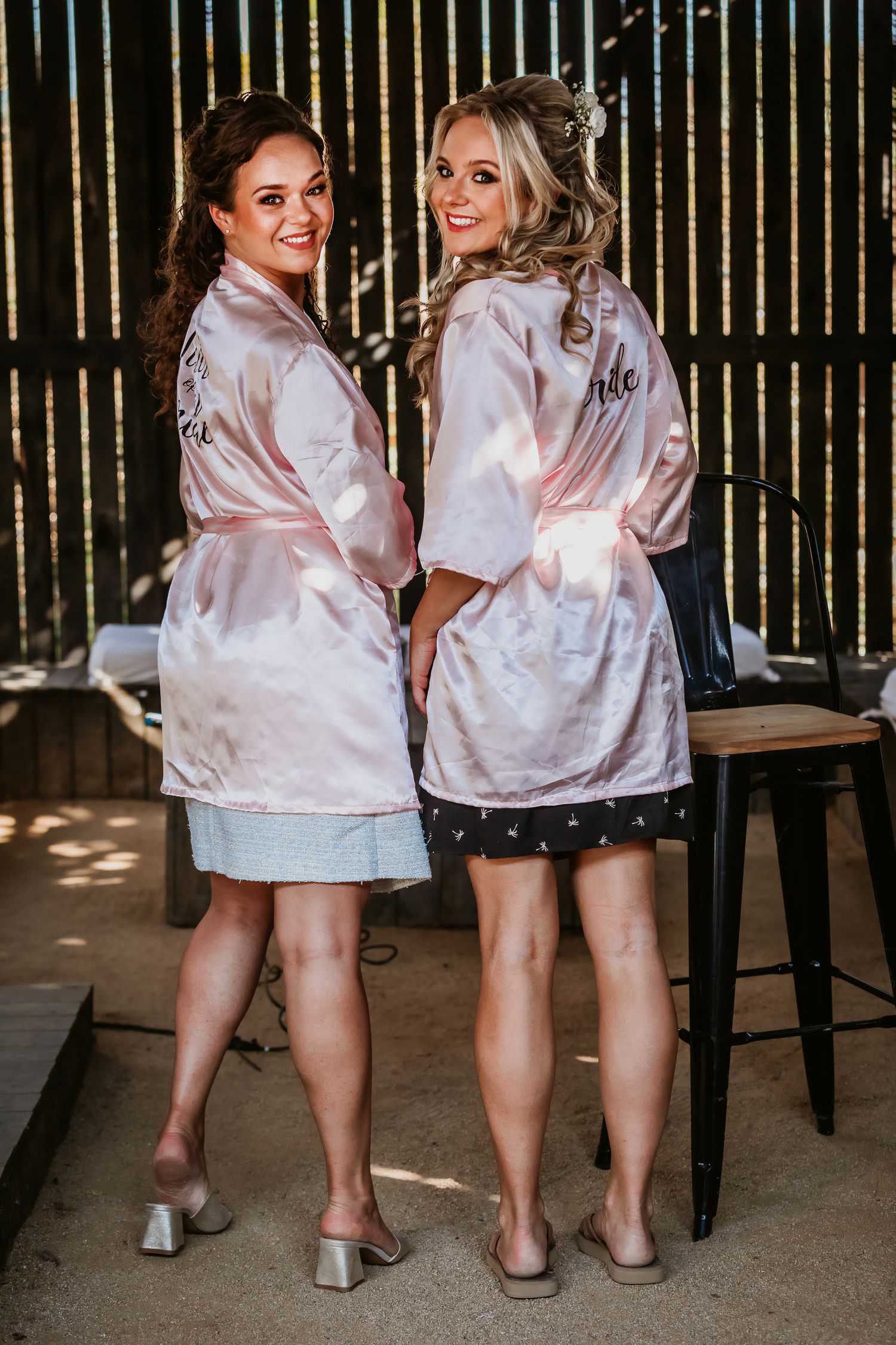 Bride and bridesmaid in pink satin robes smiling over shoulders at wedding preparation.