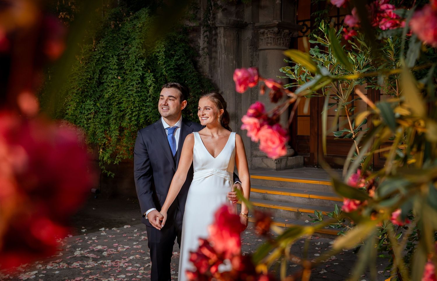 Newlywed couple holds hands surrounded by vibrant pink flowers in romantic garden setting.