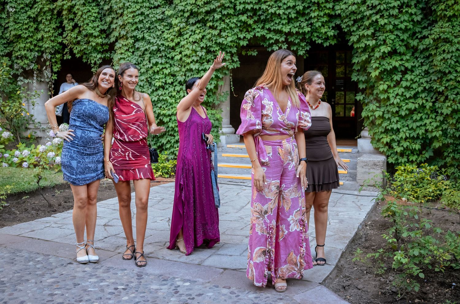 Group of women in colorful dresses posing in front of ivy-covered wall outdoors.