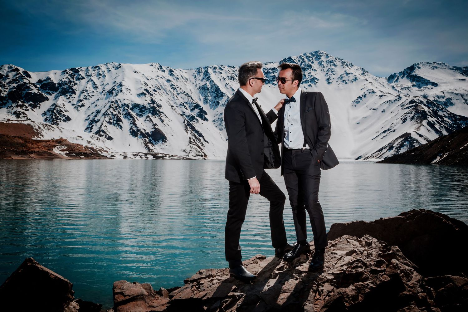 Same-sex couple in matching dark suits stand together at a scenic mountain lake during their wedding.