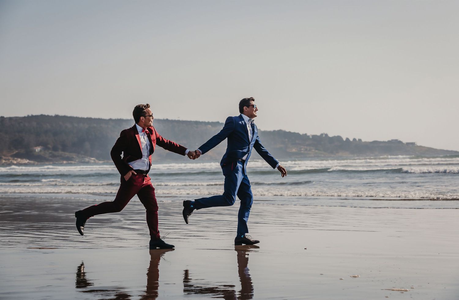 Two grooms in colorful suits joyfully run hand-in-hand along a sunlit beach shoreline.