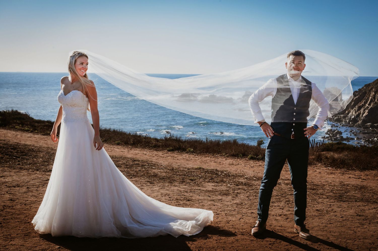 Bride and groom stand apart as her long veil billows dramatically in the wind against a stunning ocean cliff.