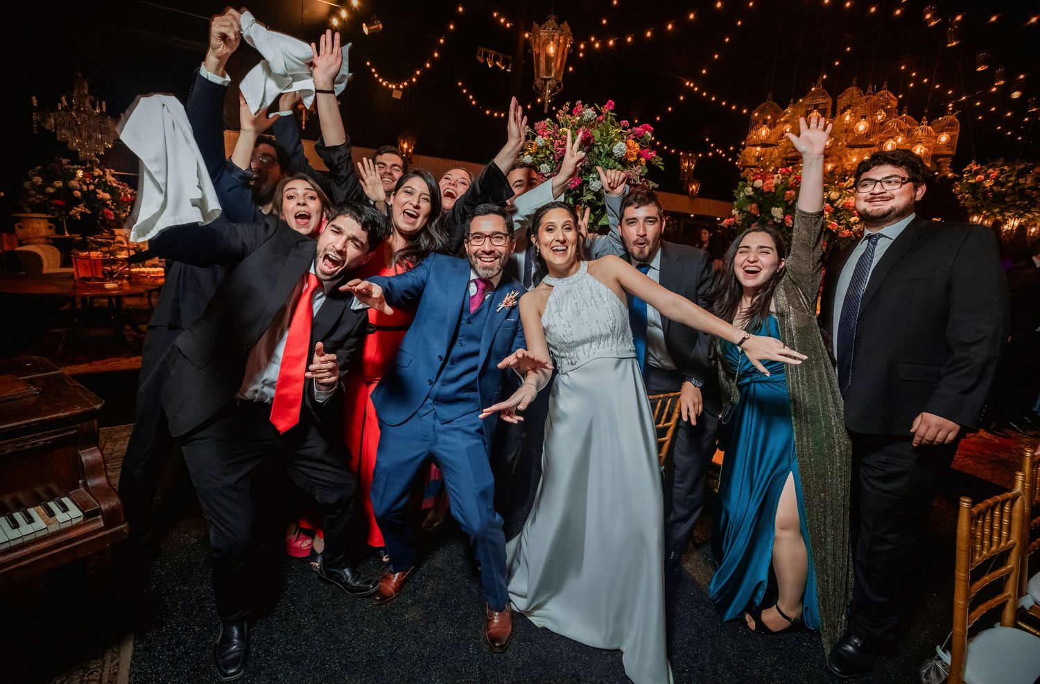 Excited wedding party poses with bride and groom under string lights at festive reception.