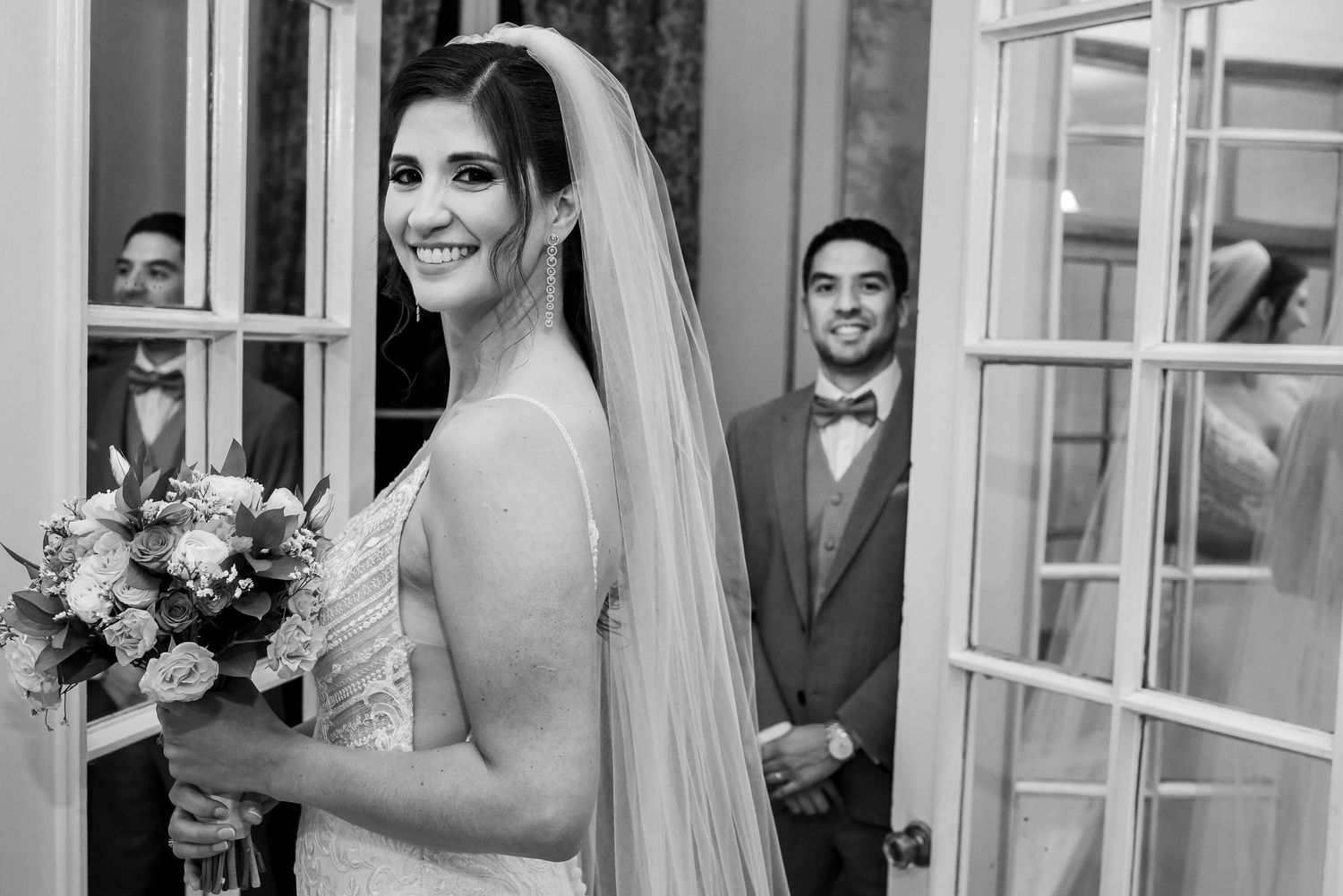 Bride in wedding dress and veil smiles holding bouquet while groom in suit stands in doorway, black and white photo.