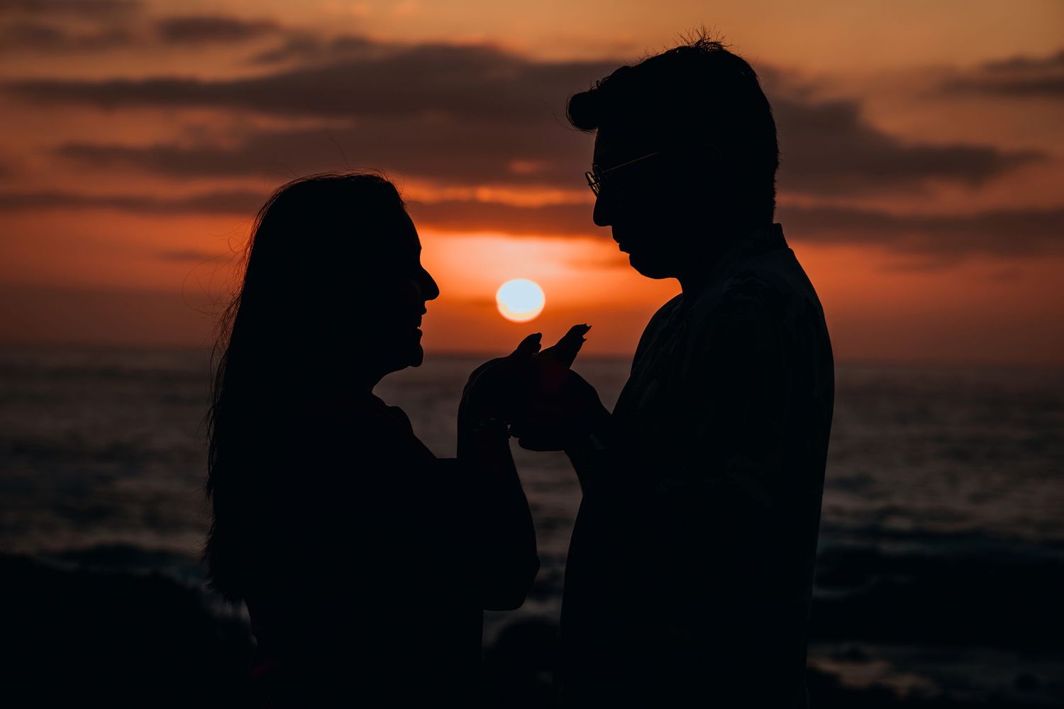 Silhouette of a couple facing each other at sunset by the ocean, with a vibrant red and orange sky.