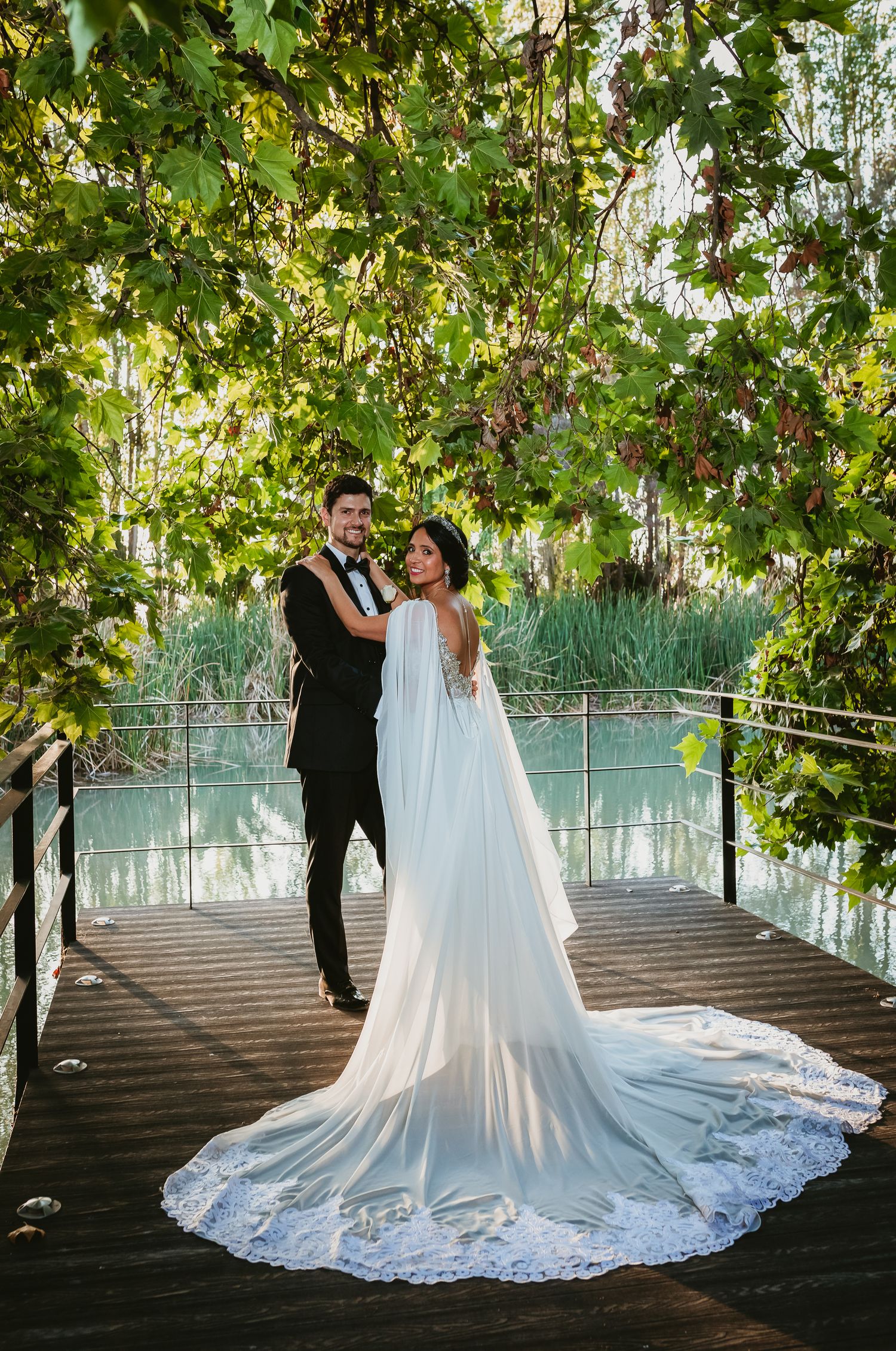 Bride and groom posing on a wooden deck surrounded by lush green vines near a tranquil pond.