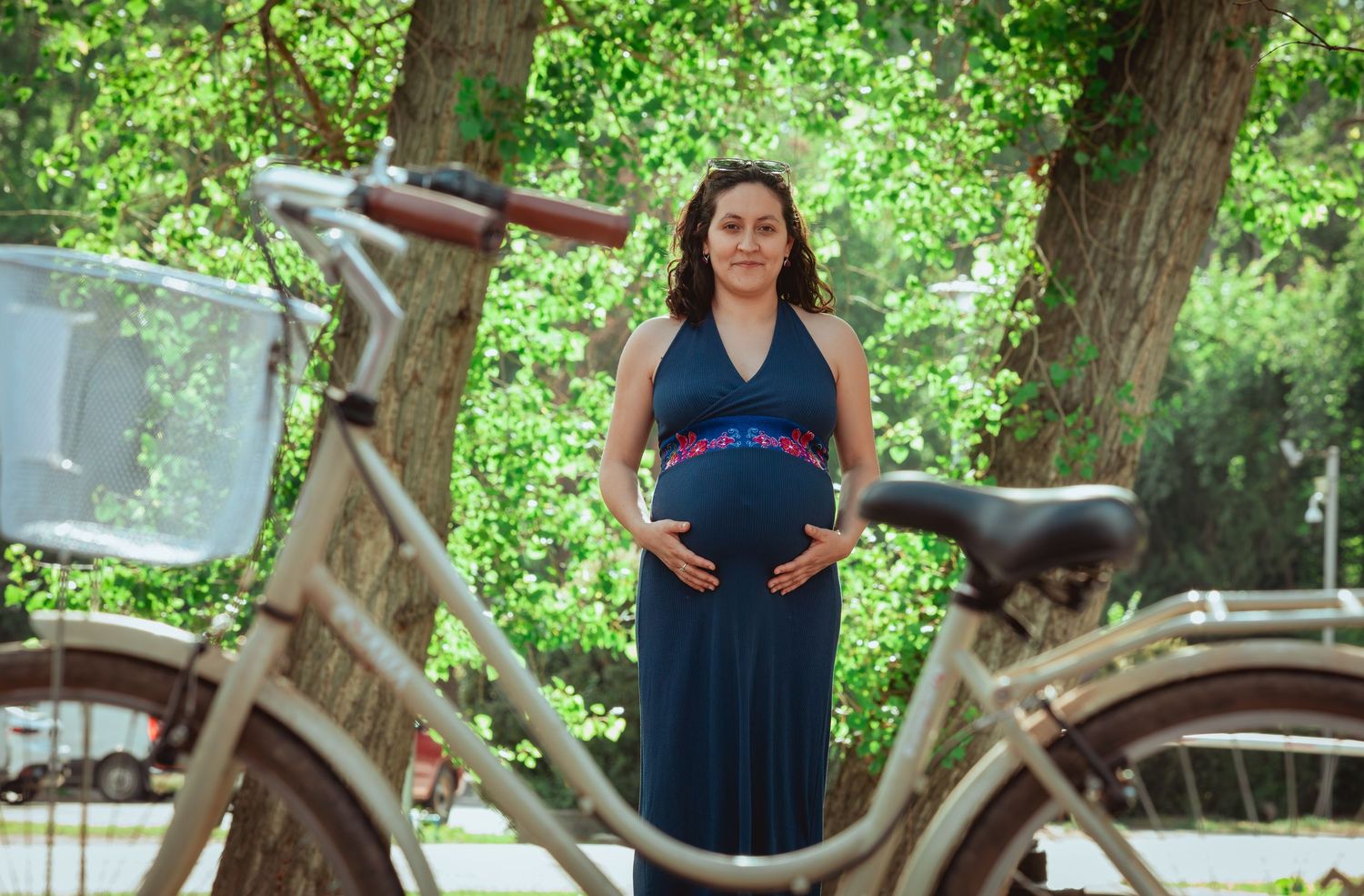 Pregnant woman in navy blue dress holding baby bump surrounded by bicycles in a lush green park.