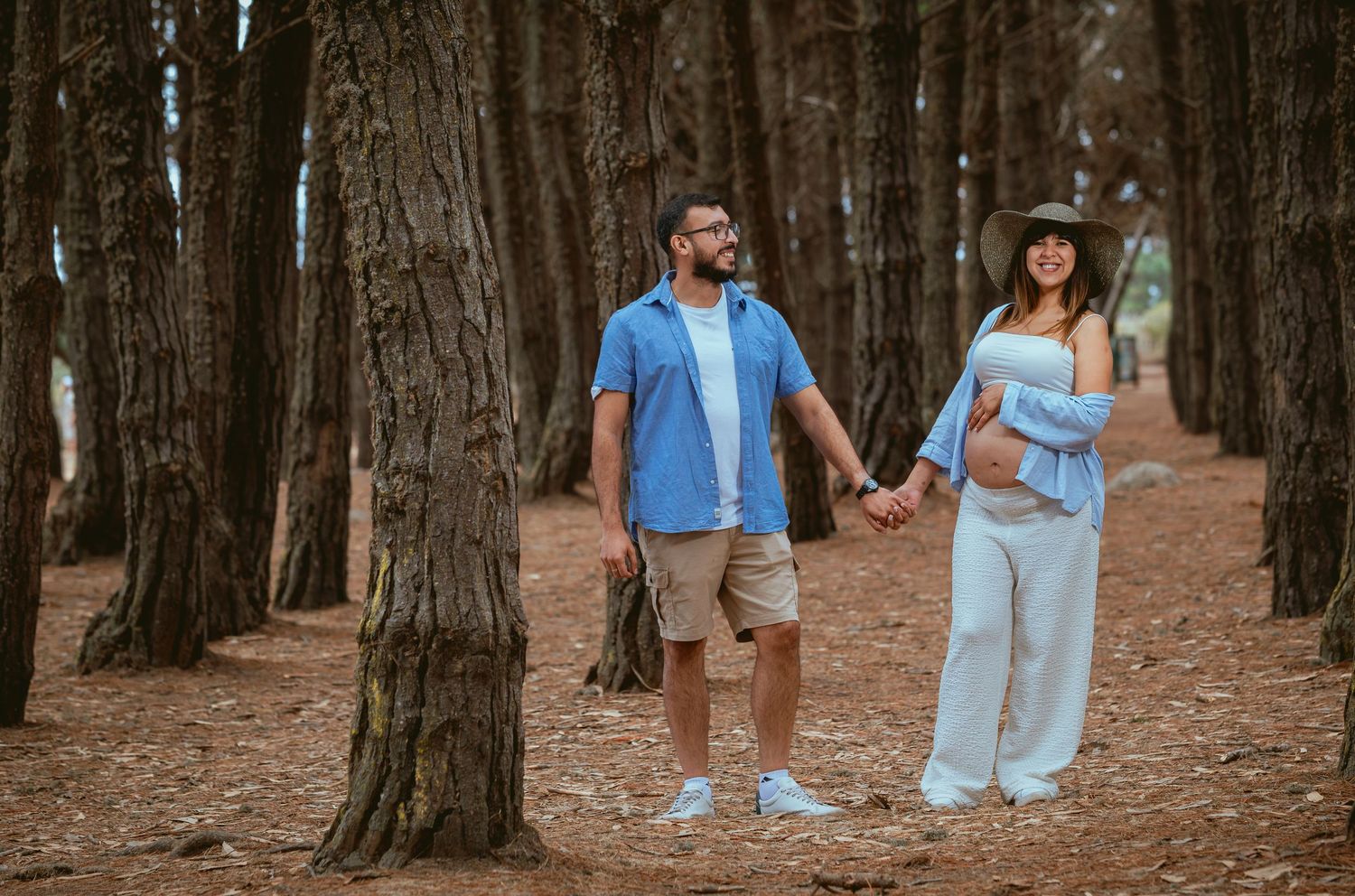 Expecting couple holding hands among tall pine trees outdoors.