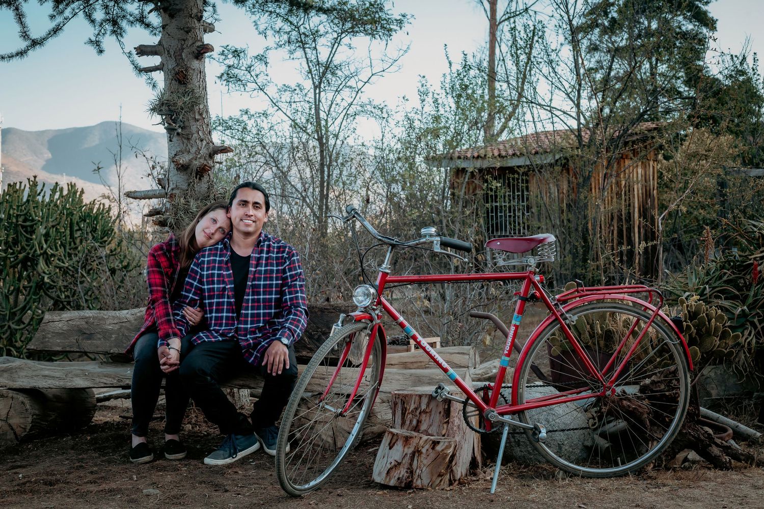 A couple sits together on a wooden bench beside a vintage red bicycle in a rural outdoor setting.