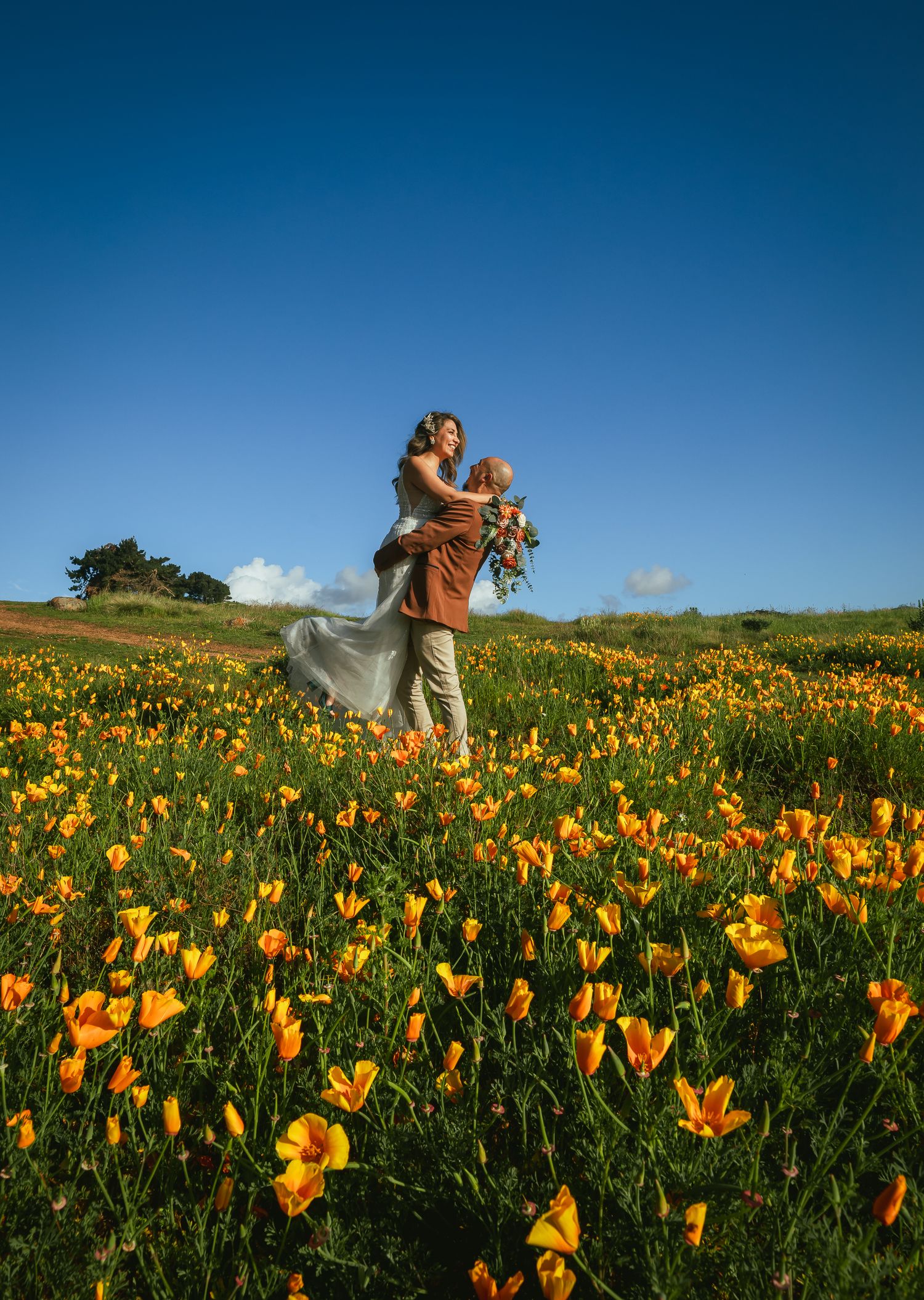 Couple embracing in a vast field of orange California poppies under a bright blue sky, the bride in a flowing white gown holding a bouquet.