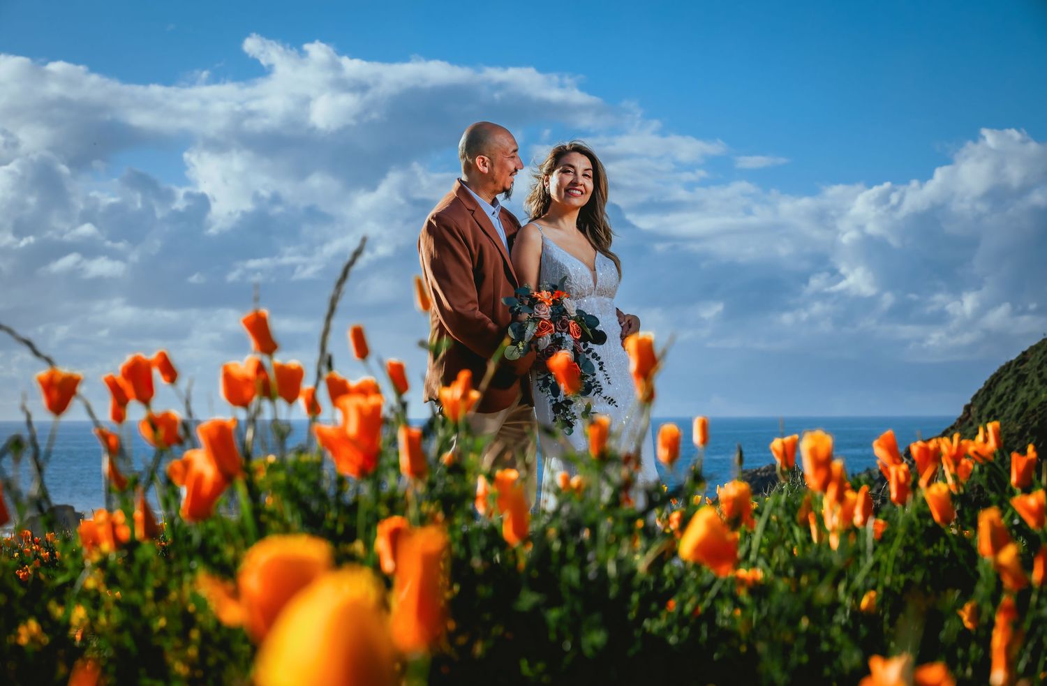 Couple embracing among bright orange poppies on a coastal hillside with the ocean and dramatic blue sky with white clouds behind them.