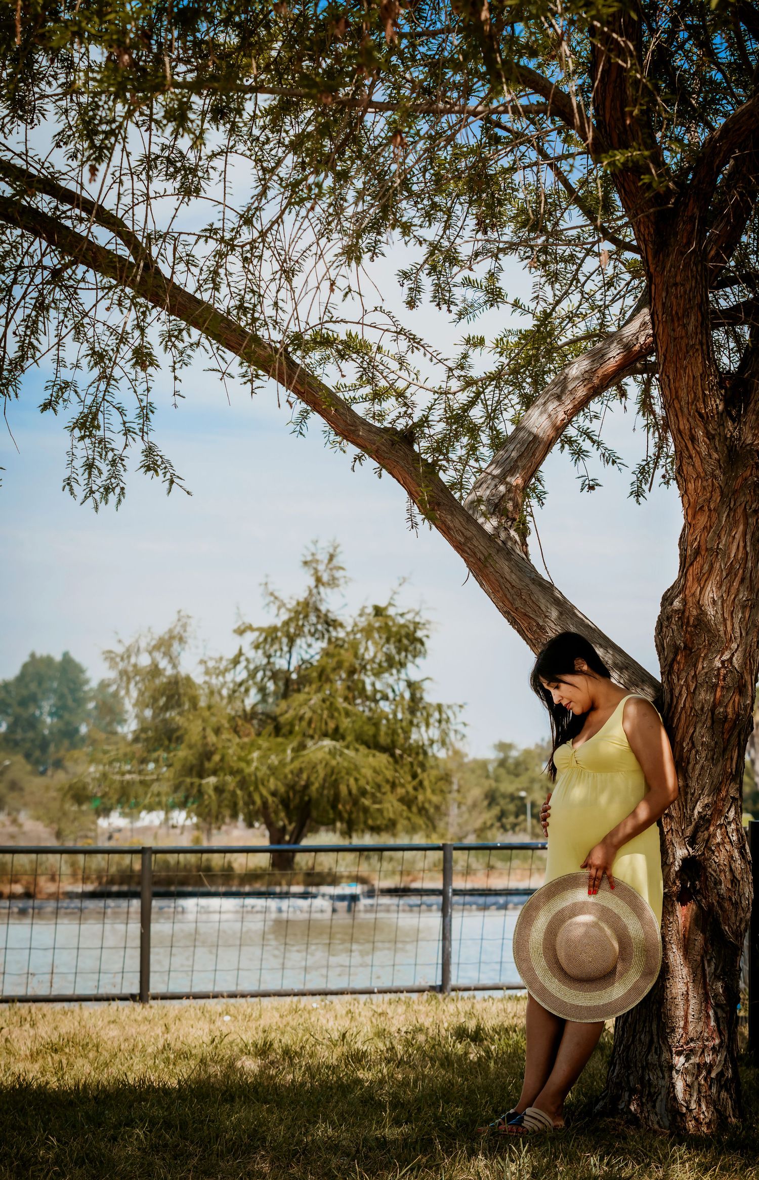Pregnant woman in yellow dress leaning against a tree by a river, holding a sun hat on a sunny day.