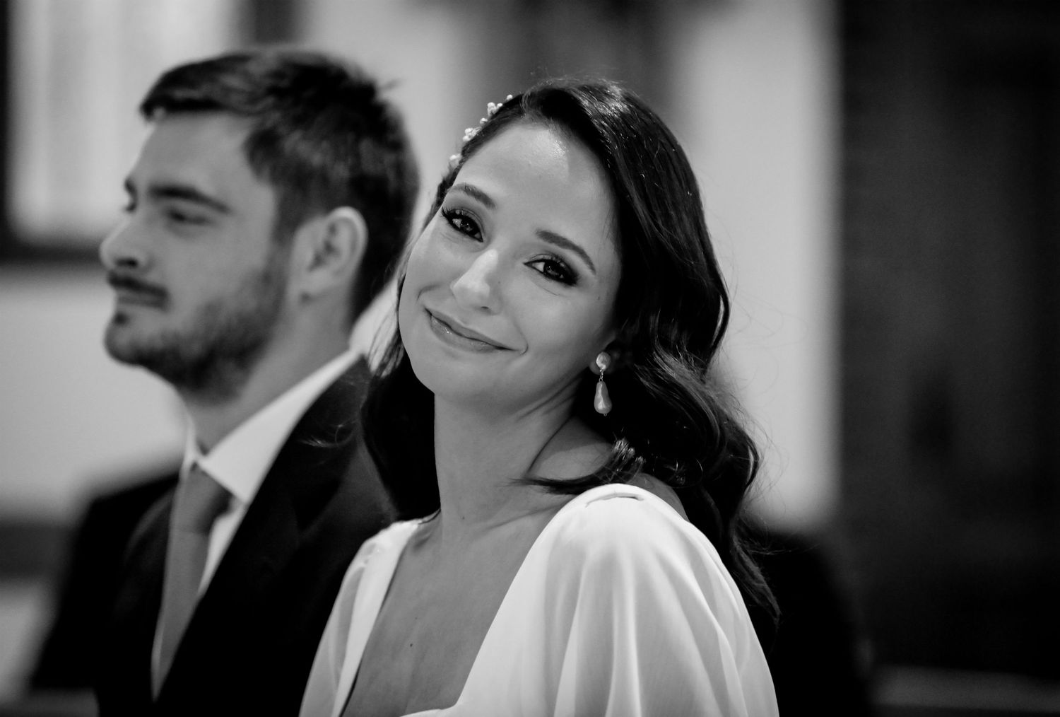 Black and white wedding photo of a smiling bride in white dress with groom in background at ceremony.