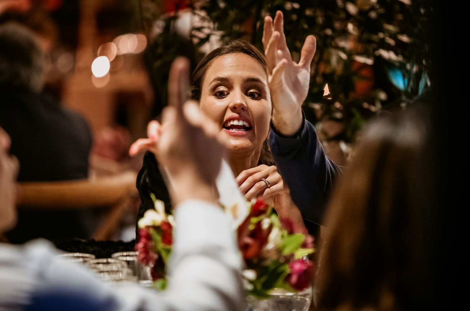 Woman smiling and gesturing with raised hands at a festive dinner party table with floral decorations.