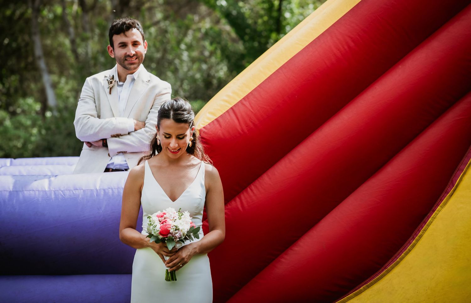 Bride in white dress holding bouquet stands before colorful inflatable slide as groom in suit smiles behind her.