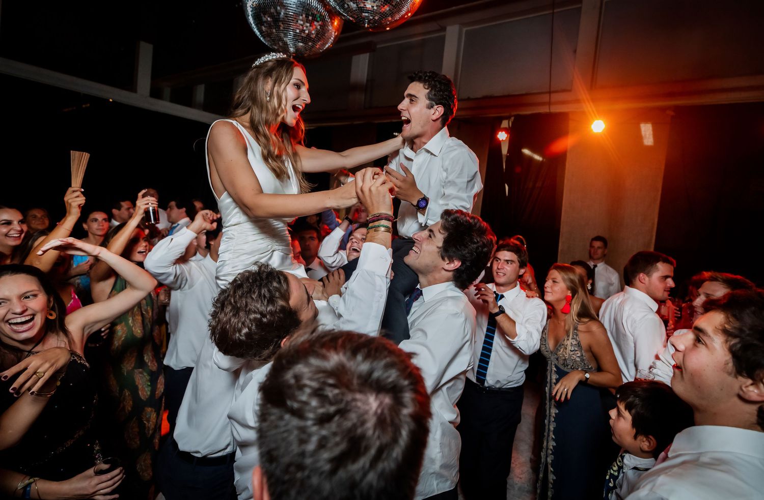Bride and groom lifted by wedding guests while holding hands above an energetic crowd at reception.