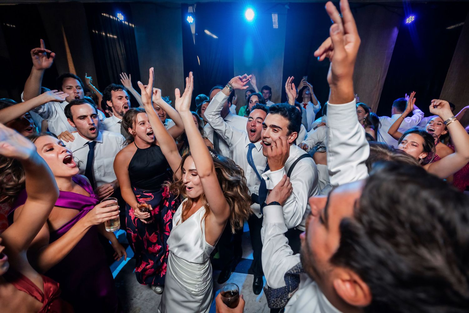 Bride and wedding guests dancing and cheering with raised arms under blue lights at reception.