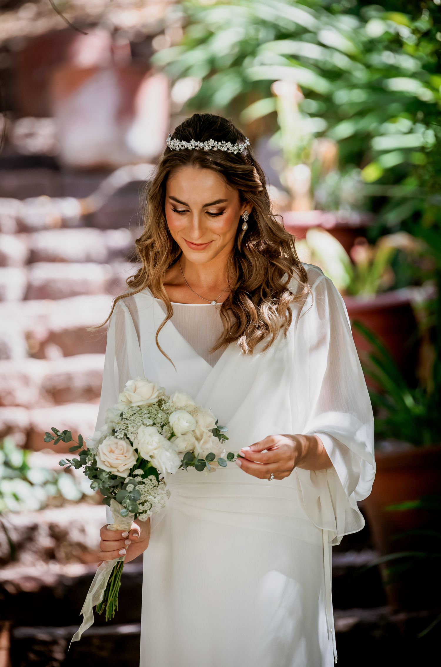Elegant bride in white gown holding bouquet of white roses with greenery, wearing crystal hair crown.