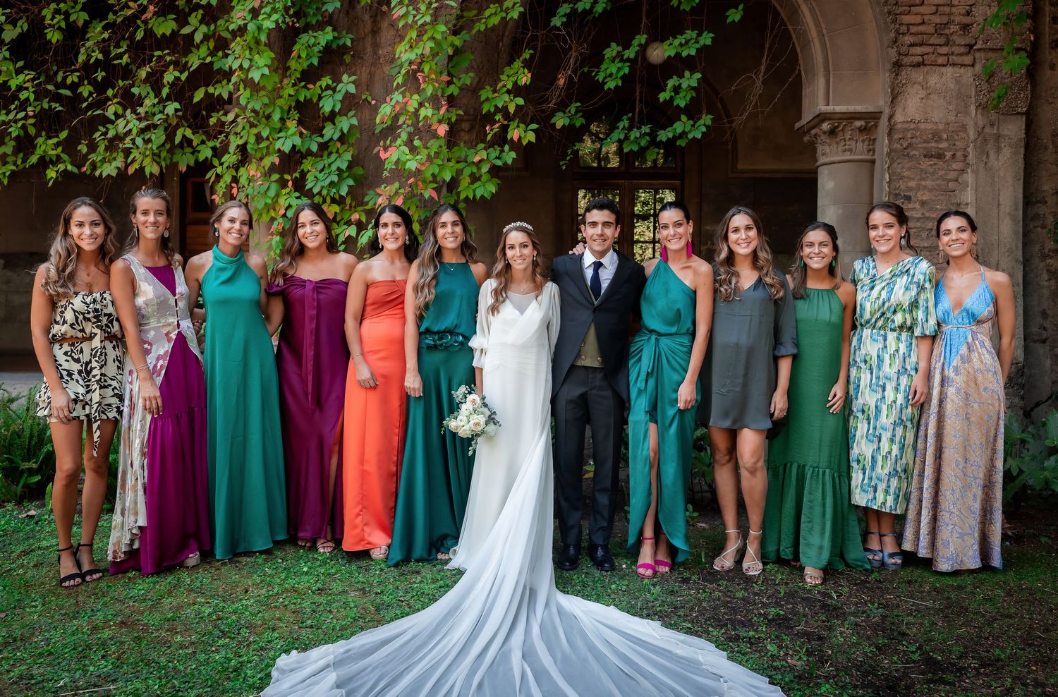 Bride and groom pose with bridesmaids in colorful teal and jewel-toned gowns outdoors.