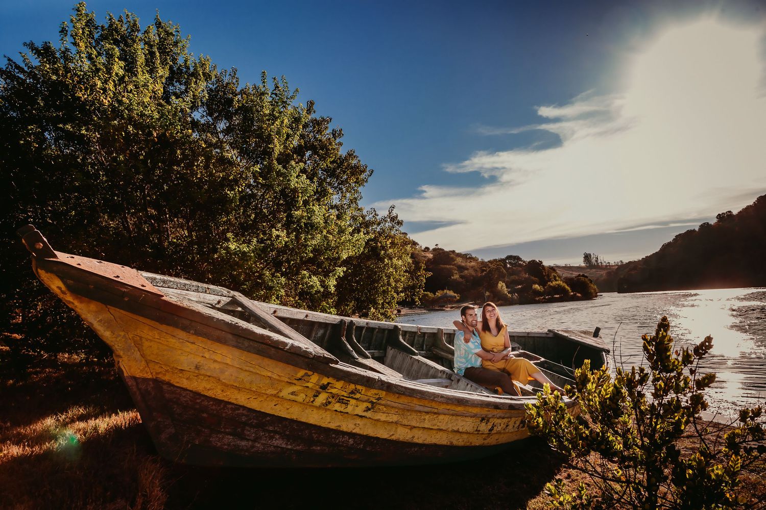 Couple sitting together in a rustic wooden boat by a sunlit riverbank with lush trees.