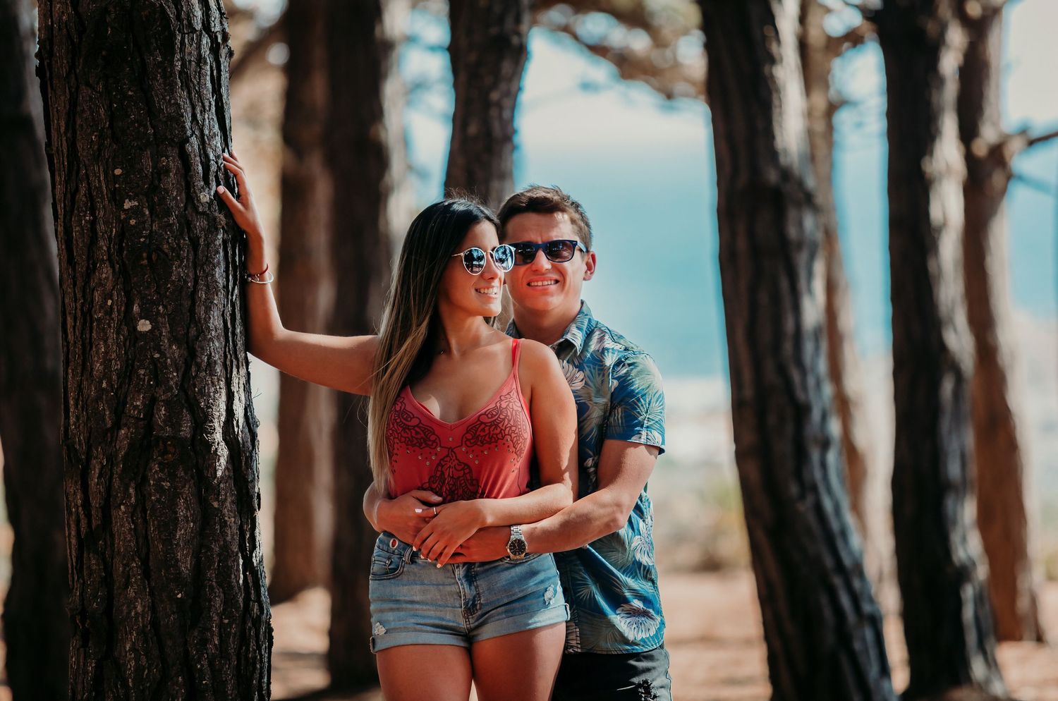 Couple embracing by a tree trunk in a pine forest, both wearing sunglasses on a sunny day.