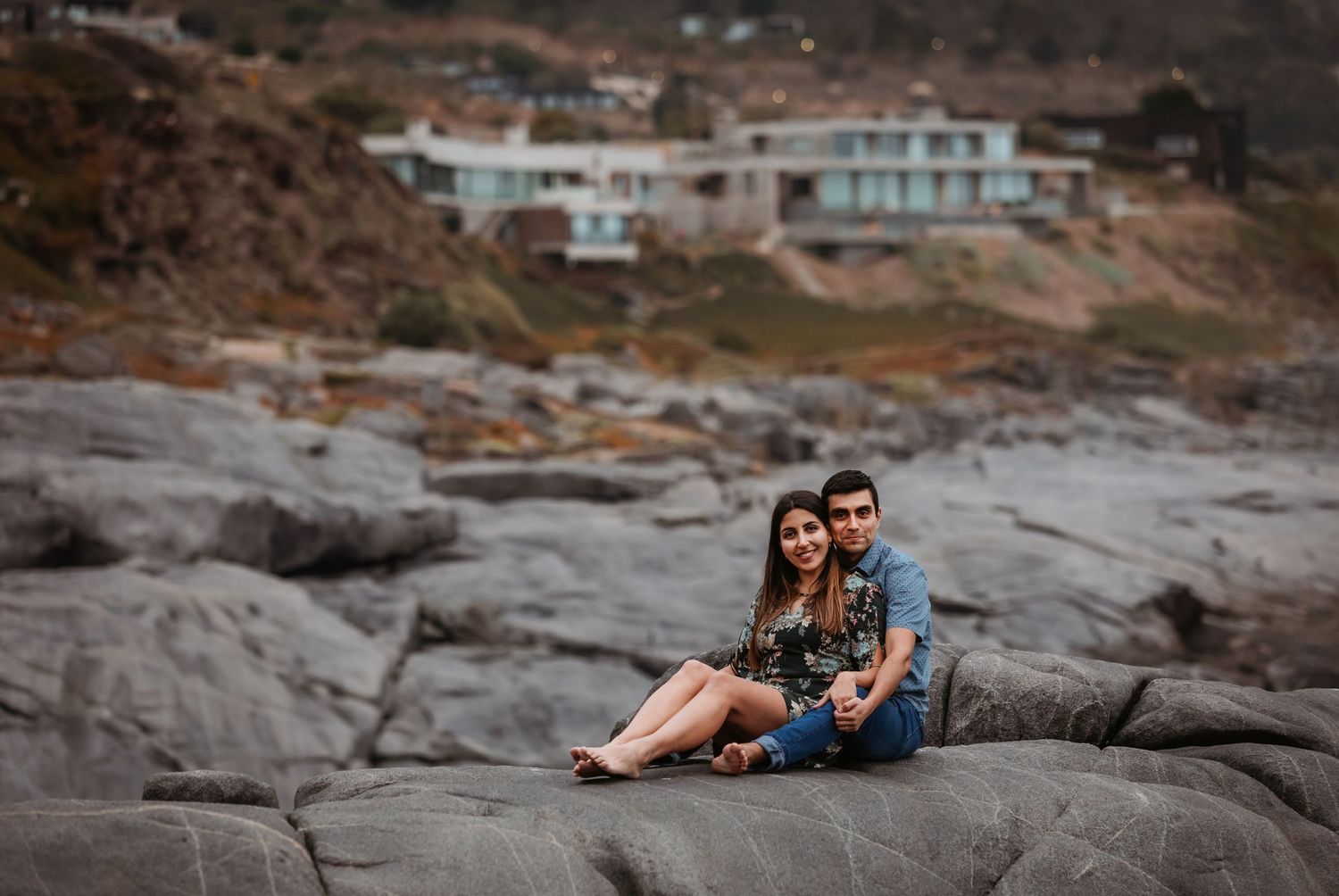 Couple sitting closely together on seaside rocks with coastal buildings visible in the background.