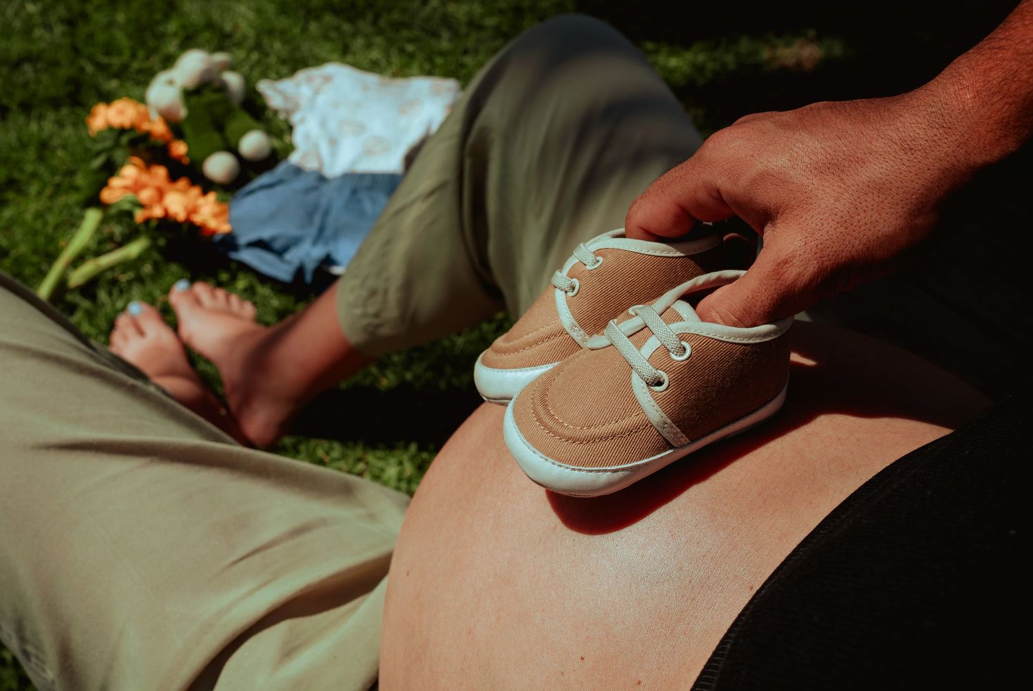 Close-up of tiny baby shoes resting on pregnant belly with flowers and baby clothes on grass nearby.