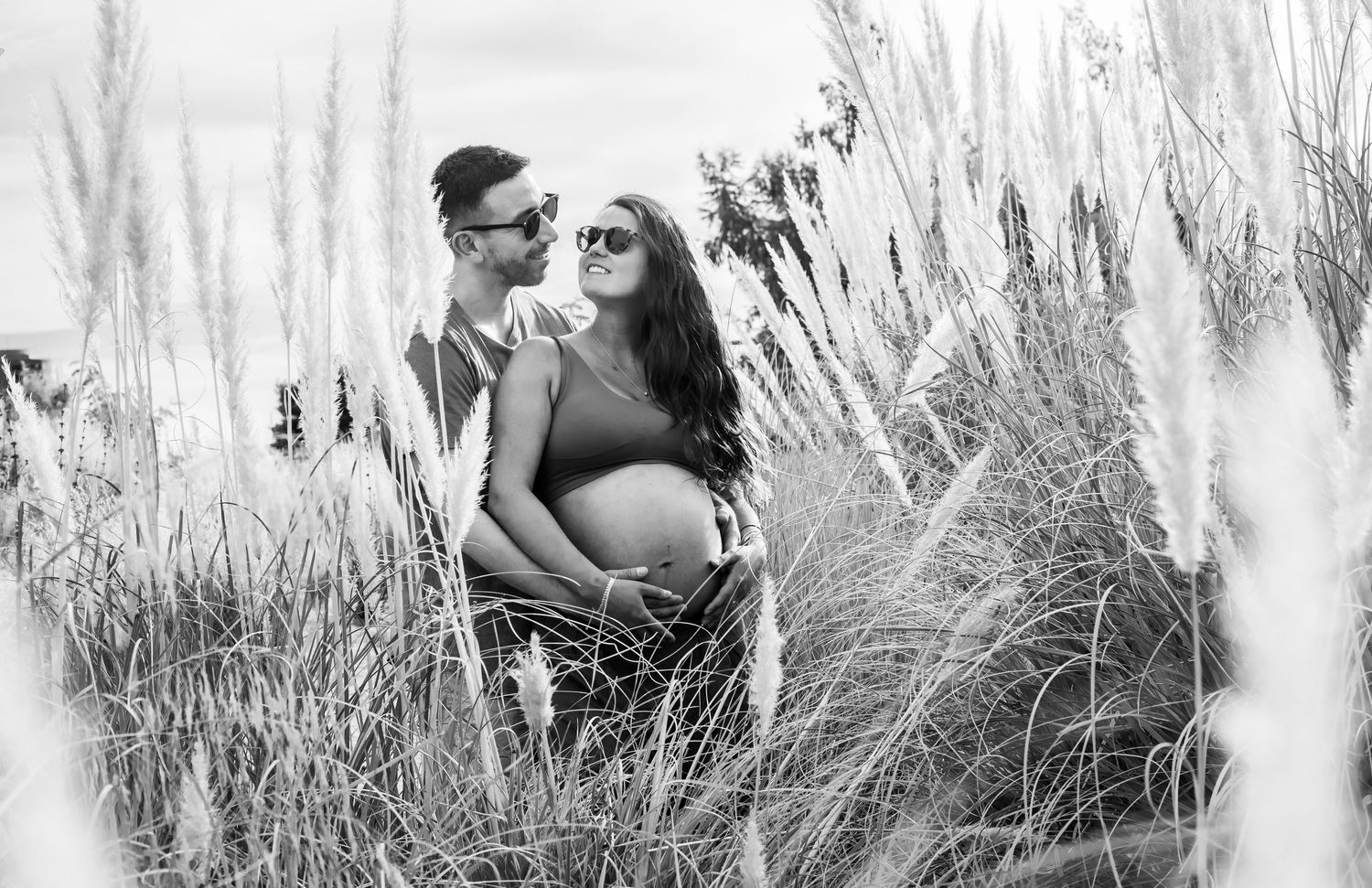 Black and white photo of couple embracing in tall grass, woman with pregnant belly.