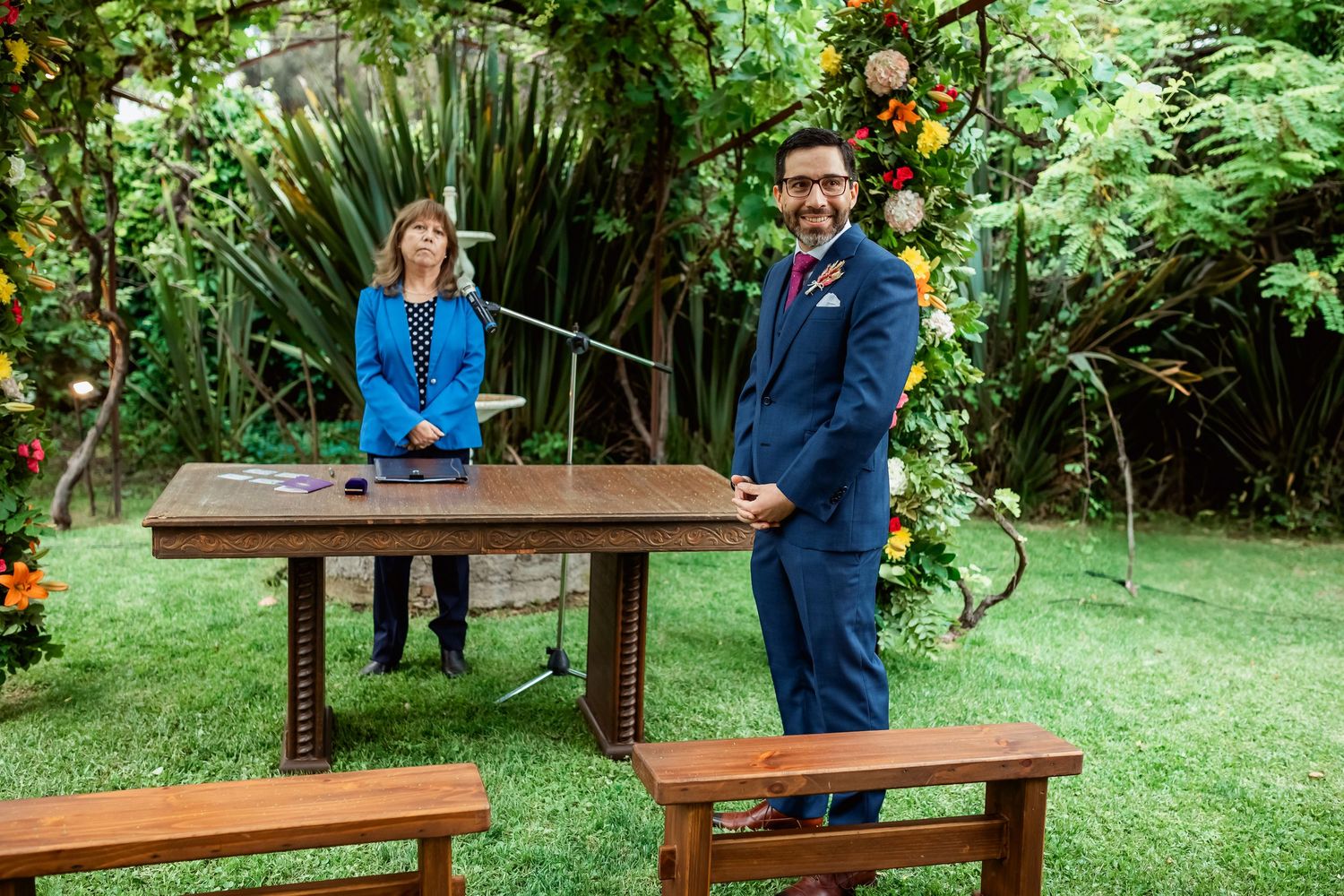 Groom in navy suit stands beside officiant at wooden ceremony table in lush green outdoor garden setting.