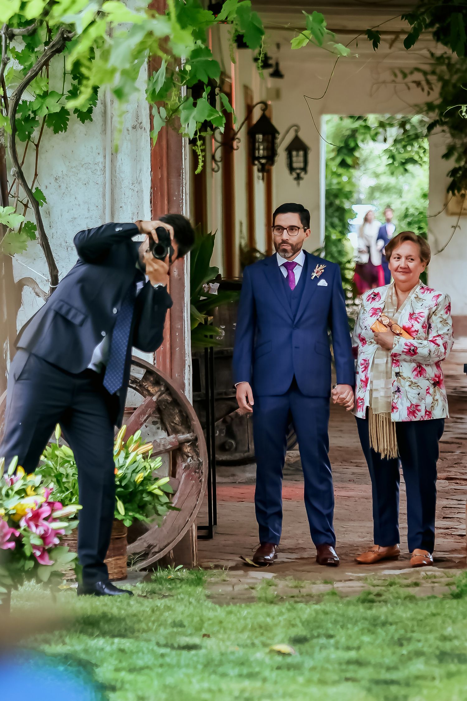 Wedding photographer captures groom in blue suit with older woman in floral dress at rustic outdoor venue.